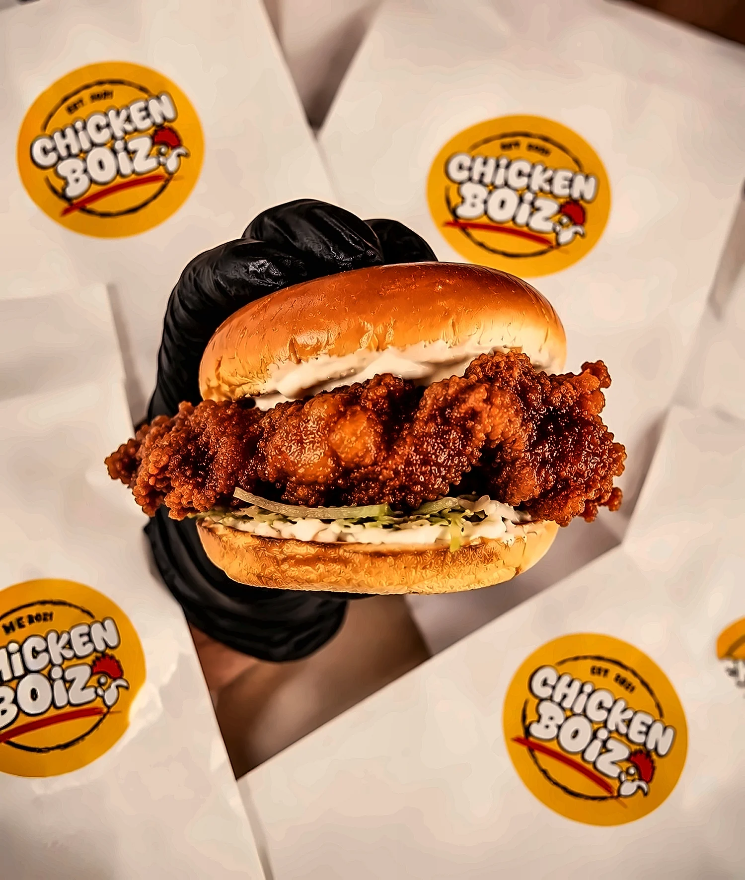 A crispy chicken burger in a black glove, on Chicken Boiz branded paper covered in yellow logo stickers